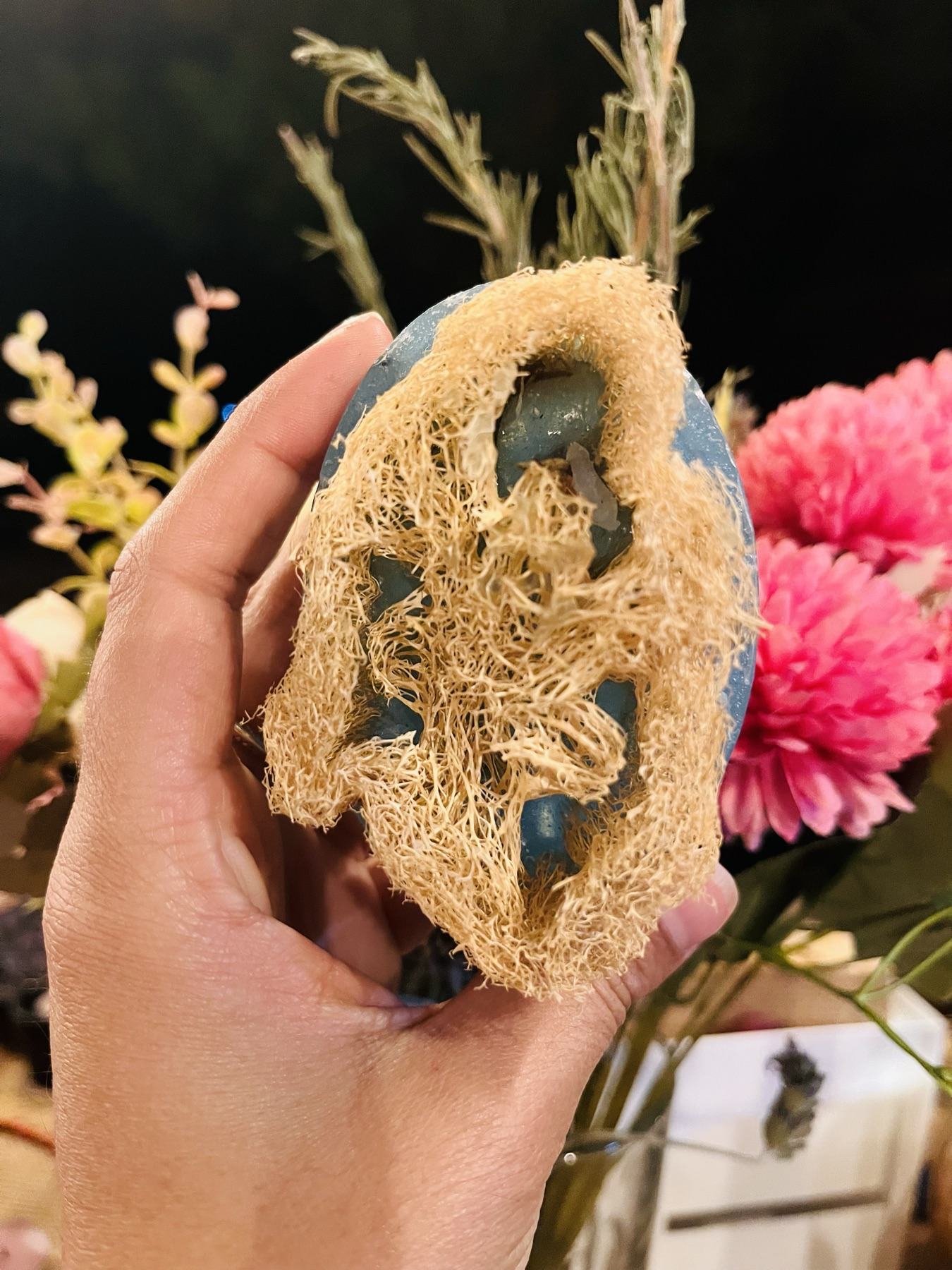 Jabón artesanal con esponja de lufa sostenido en la mano, entre flores y ramas secas